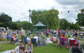 Groups of people, in the meadow, bandstand in background.