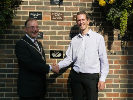 Mayor and man shake hands in front of brick wall after unveiling a commemorative plaque.