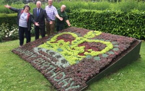 Three males and one female standing behind a flower bed in the design of a crown and with the words happy birthday.