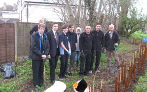 Four boys in school uniform with five adults. Standing in front of newly planted hedge.