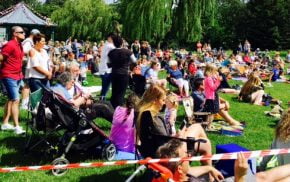 Large crowd of people sit and stand on grass to watch dance festival. Blue sky.