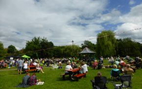 People sitting on grass and at picnic tables. Blue sky, bandstand in background.