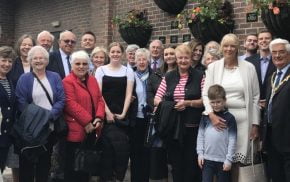 Large group of people standing in front of a wall displaying plaques and hanging baskets.