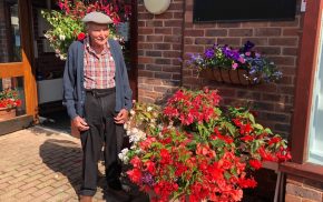 Elderly man standing next to container overflowing with red flowers and plants.