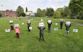 Group of people standing 2m apart on grass holding signs.