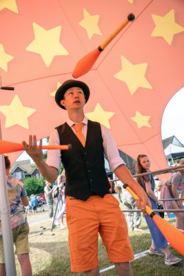 Man wearing orange shorts and black waistcoat juggling with clubs.