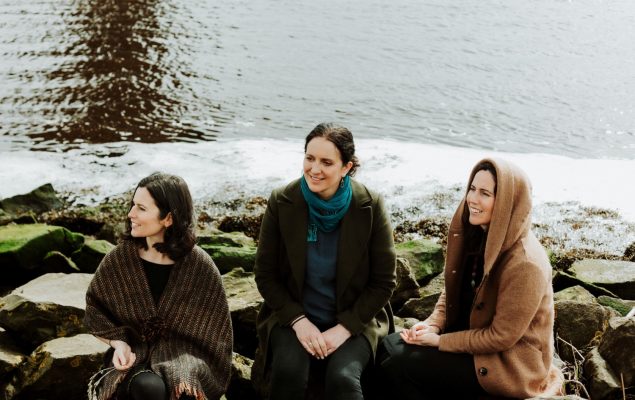 Three females sat on rocks by the sea, dressed in warm clothing and smiling.