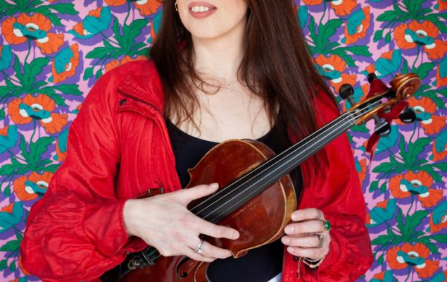 Photo of a woman stood infant of a patterned printed wall and holding an instrument