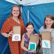 Woman and two girls showing a cross stitch sample.