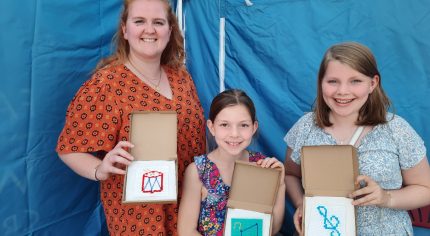 Woman and two girls showing a cross stitch sample.