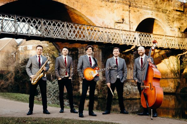 Photo of 5 men in grey suits stood in front of a bridge holding musical instruments