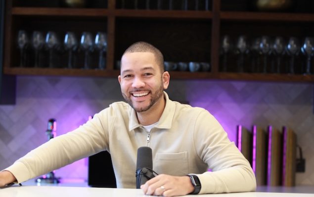 Photo of a man in a beige shirt holding a microphone and smiling at the camera